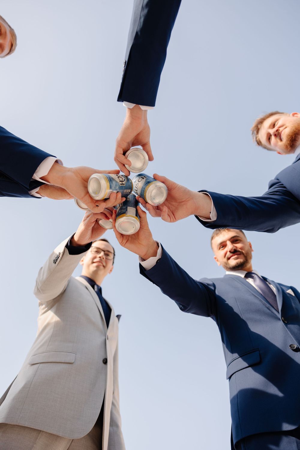 Wedding party members toast with beer cans against a bright sky background.