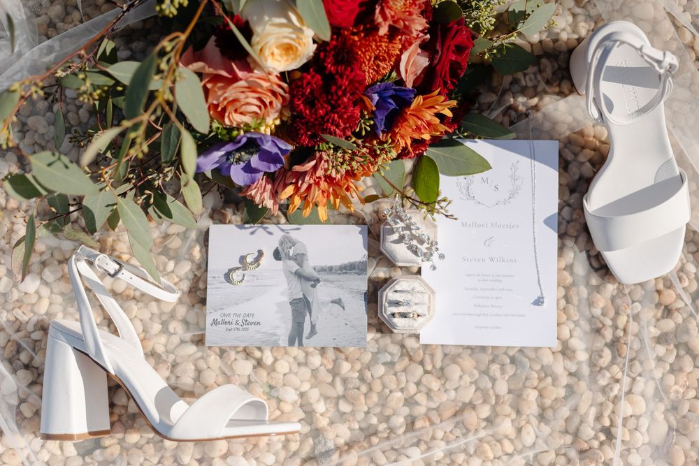 Scattered wedding details including invitations, ribbons, flowers, and white shoes arranged artistically on textured stone surface.