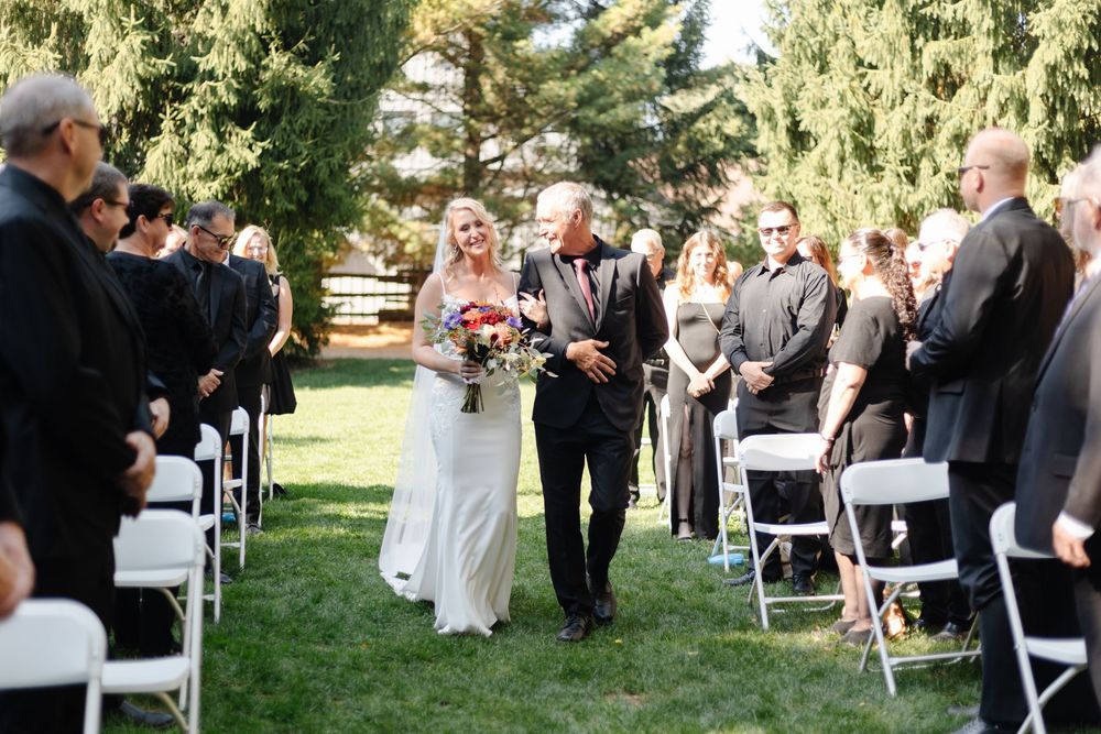 Outdoor wedding ceremony setup with white chairs and guests gathering under sunlit trees in a garden setting.