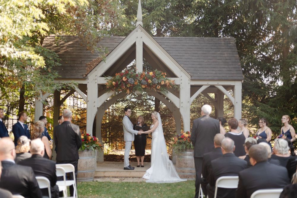 Wedding ceremony under a rustic wooden pavilion with guests seated on white chairs.