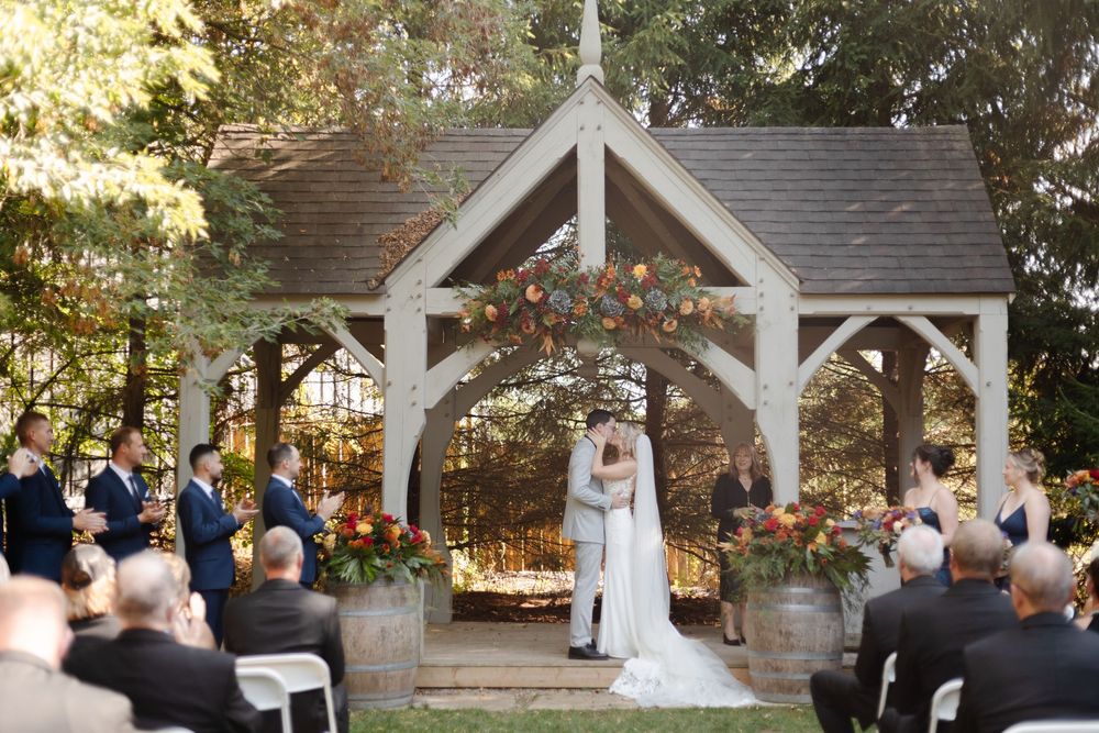 Outdoor wedding ceremony under a white wooden gazebo surrounded by trees and guests.