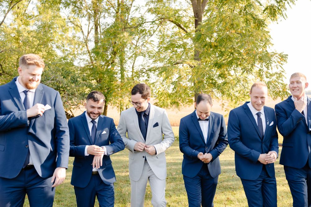 Wedding party in navy blue suits stand in a line against autumn trees at outdoor celebration.