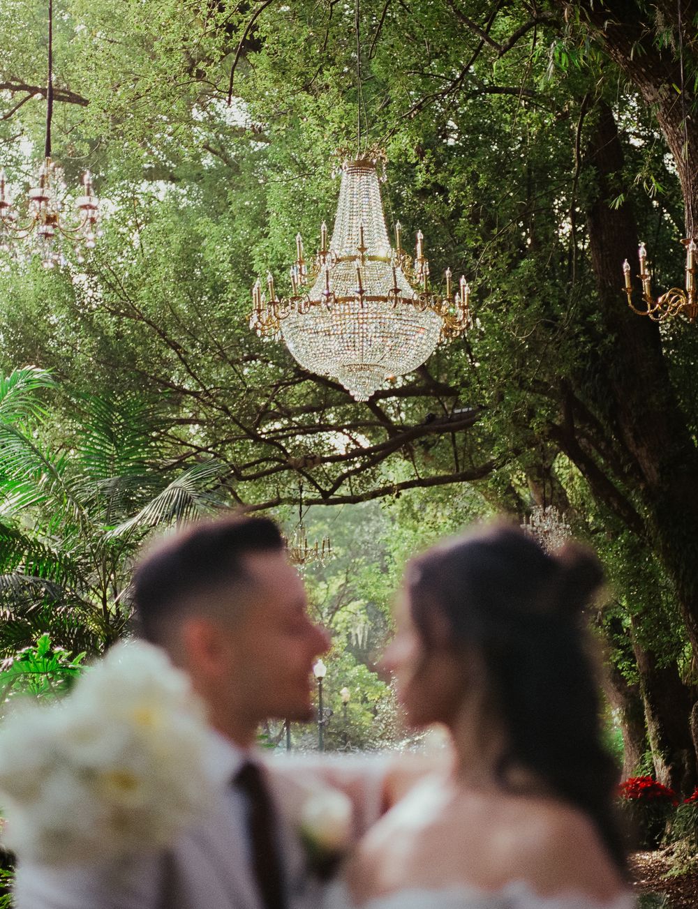 An elegant crystal chandelier hangs from tree branches in an enchanted garden setting during a romantic moment.