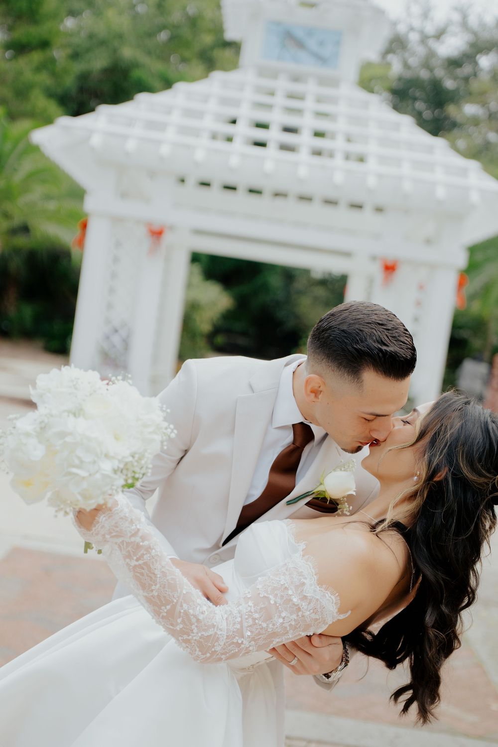 A romantic dip kiss between newlyweds in front of a white garden gazebo during their wedding celebration.