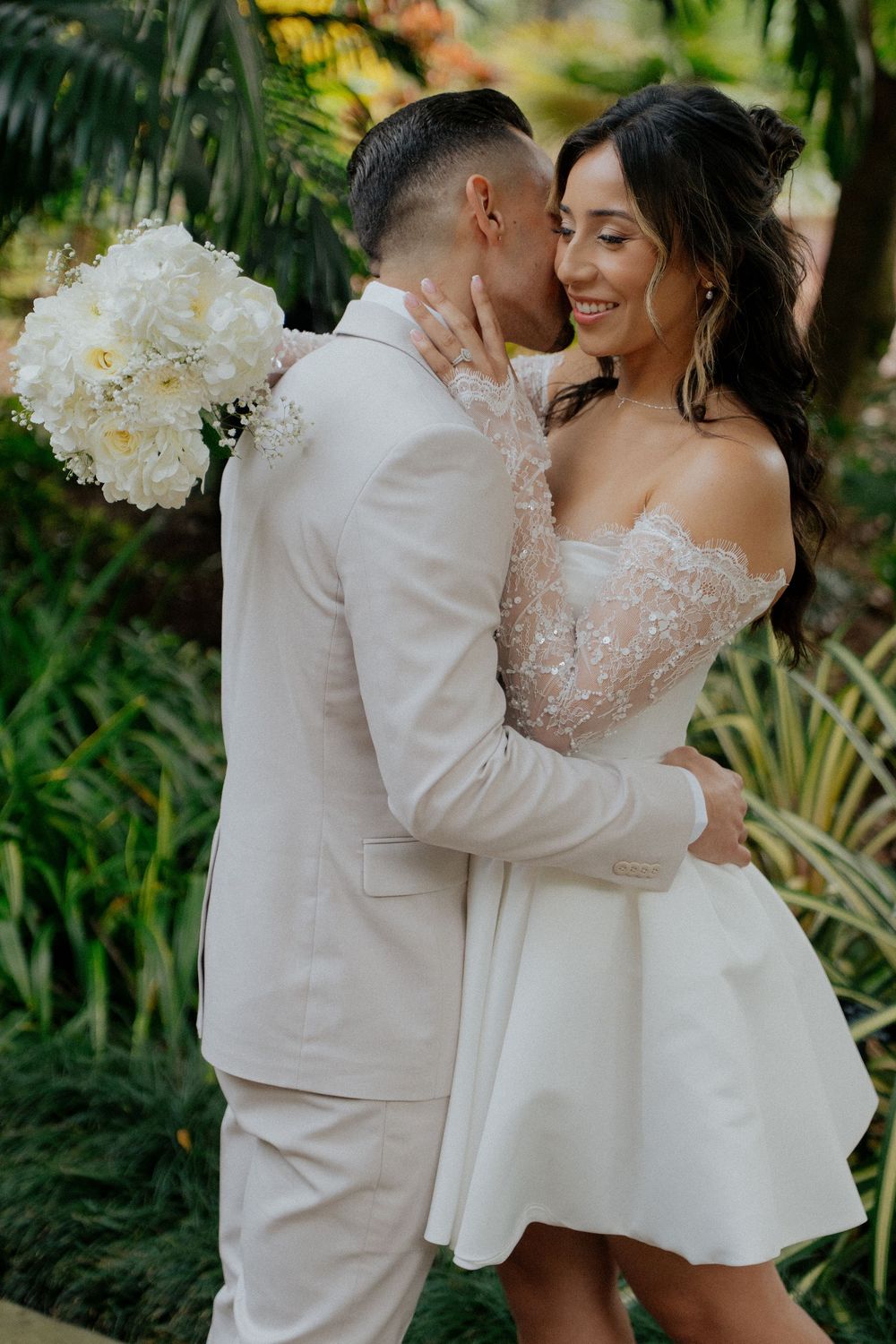 A romantic wedding couple shares intimate moments in a lush tropical garden setting with white floral bouquet.