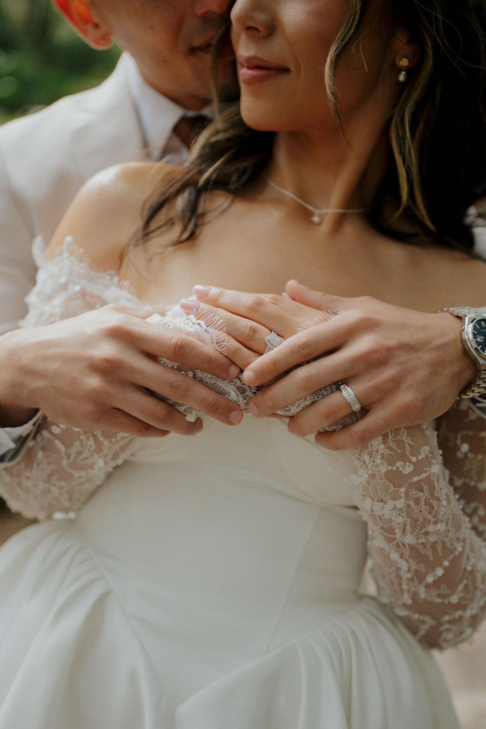 Close-up detail of hands with wedding rings against a white lace wedding dress in natural lighting.
