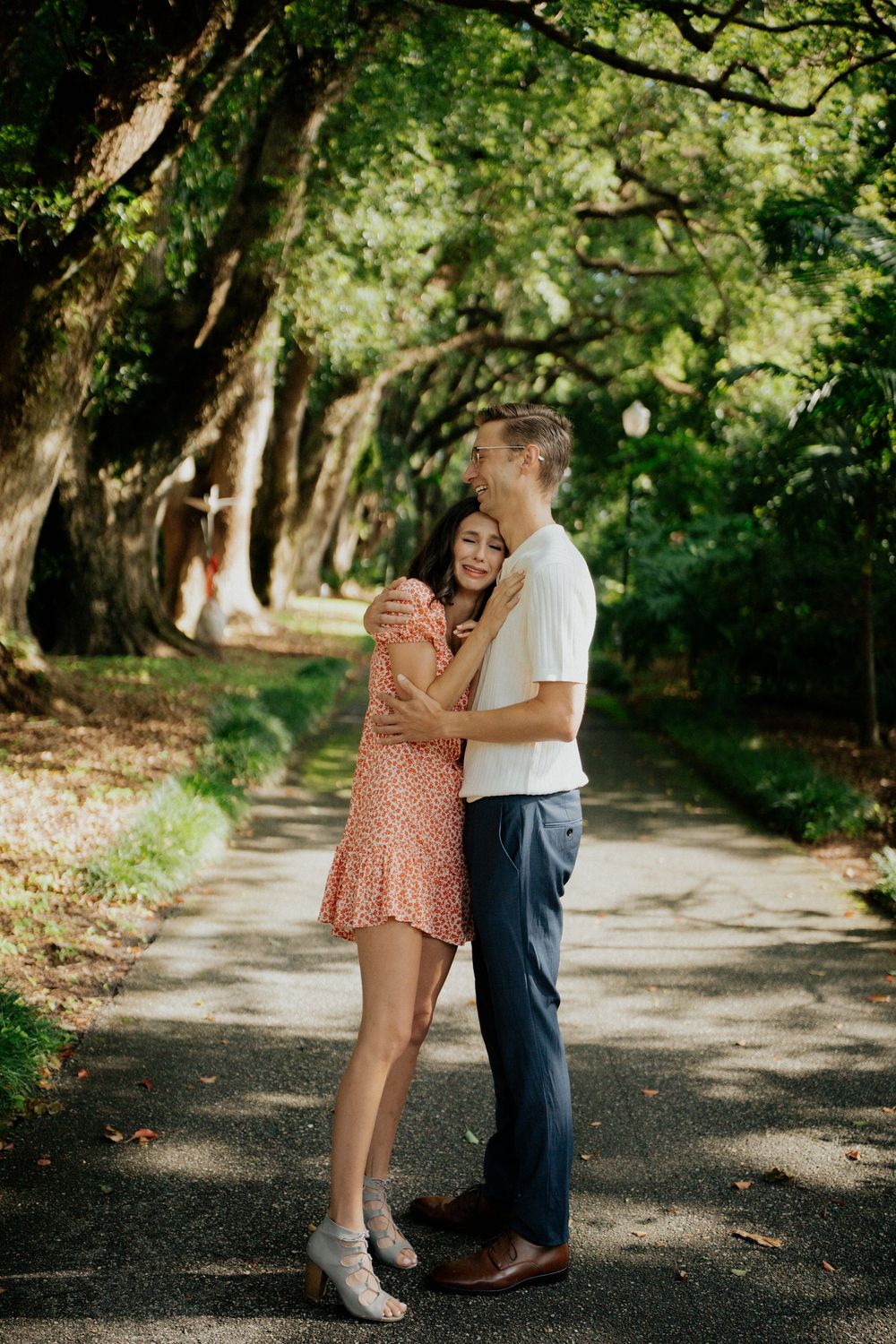 A romantic couple embraces on a tree-lined path during a summer photoshoot in a park setting.