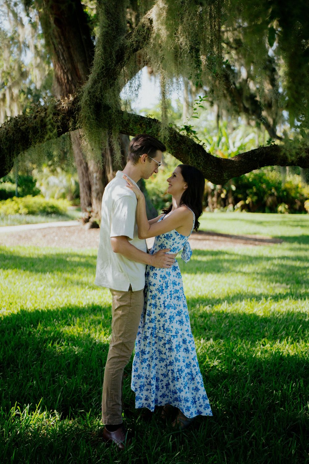 A romantic couple shares tender moments under moss-draped trees in a lush green park setting.