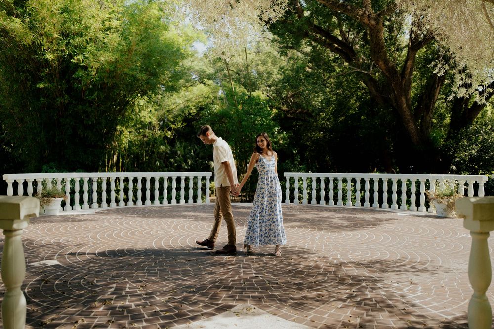 A couple stands together on a stone terrace with white balustrades surrounded by lush greenery on a sunny day.