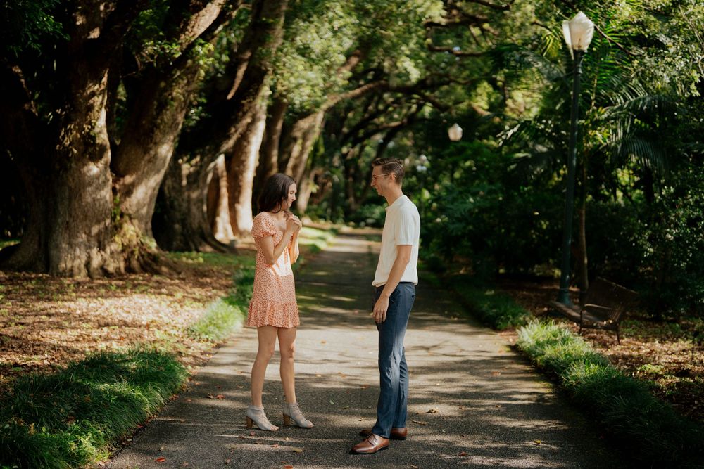 A couple walks together on a tree-lined path during a sunny summer engagement photo session.
