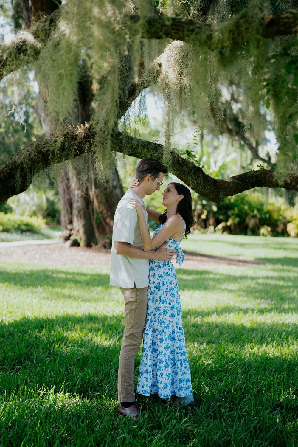 A romantic couple embraces under Spanish moss draped oak trees in a sunlit Southern garden setting.