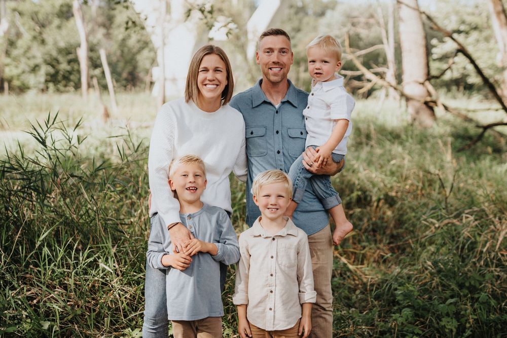 A family of five poses together outdoors in coordinated casual outfits against a natural wooded background.