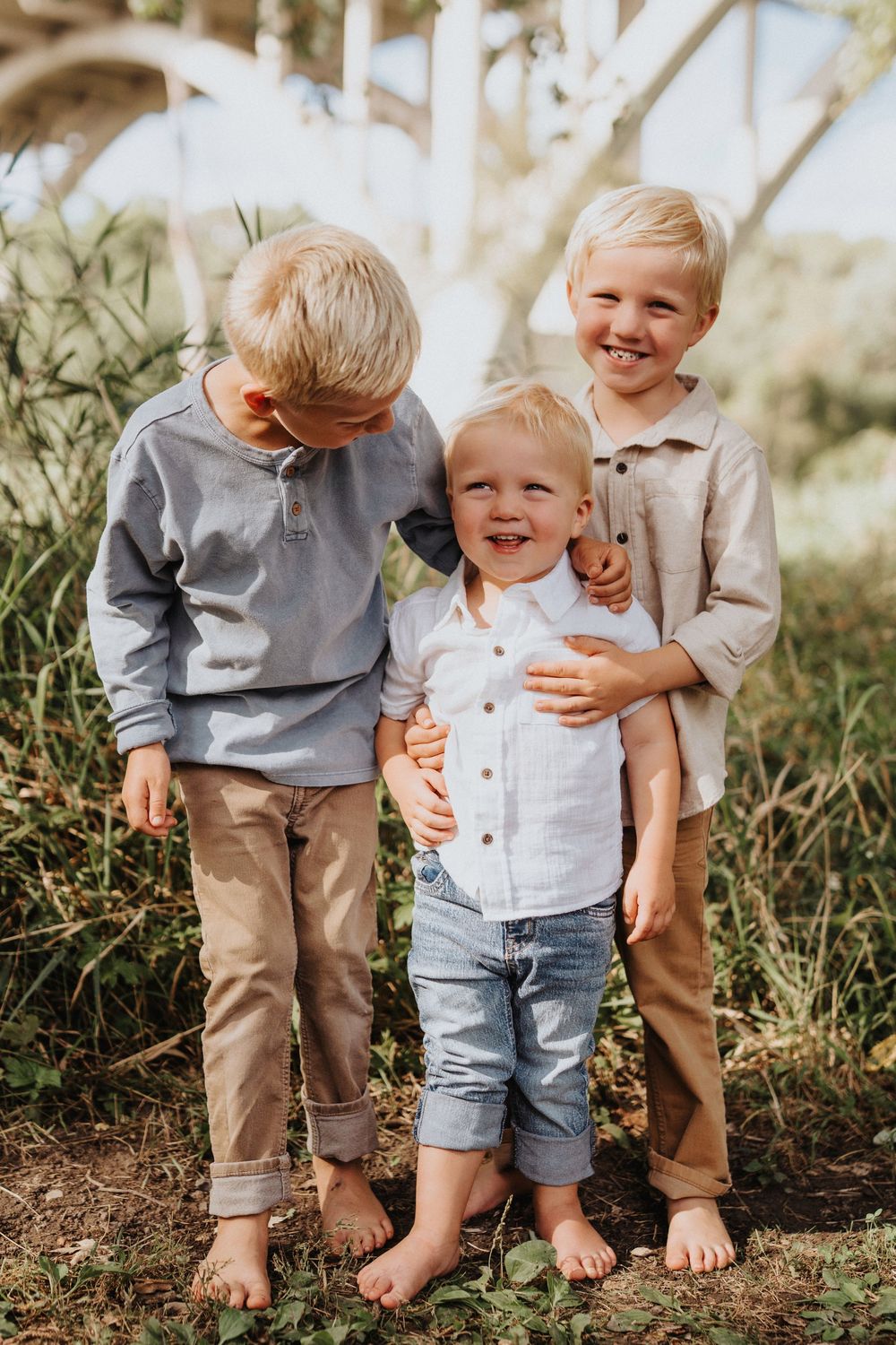 Three barefoot children in casual clothing share laughs together while posing outdoors in nature.