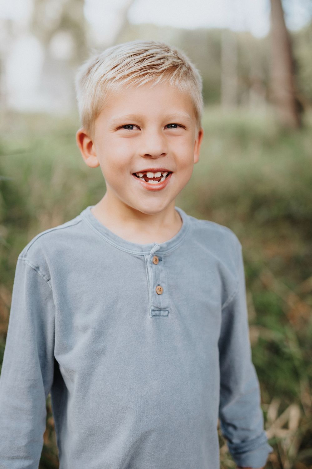 Young child wearing a gray henley shirt smiles outdoors with a missing tooth in a natural setting.
