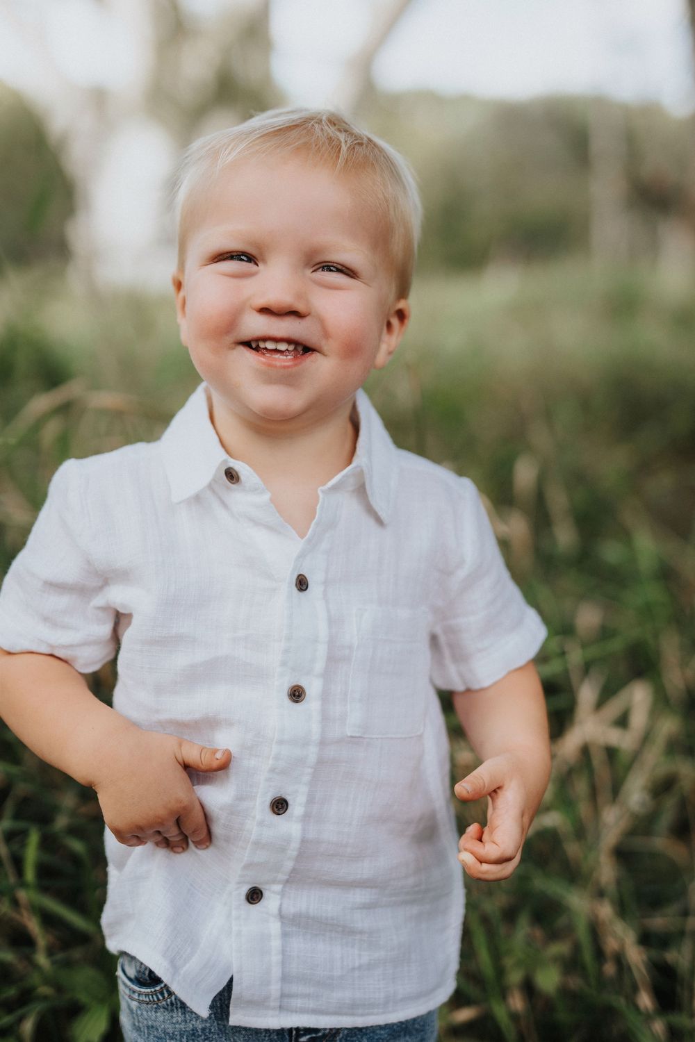 Cheerful young child in white button-down shirt smiling outdoors among tall grass.
