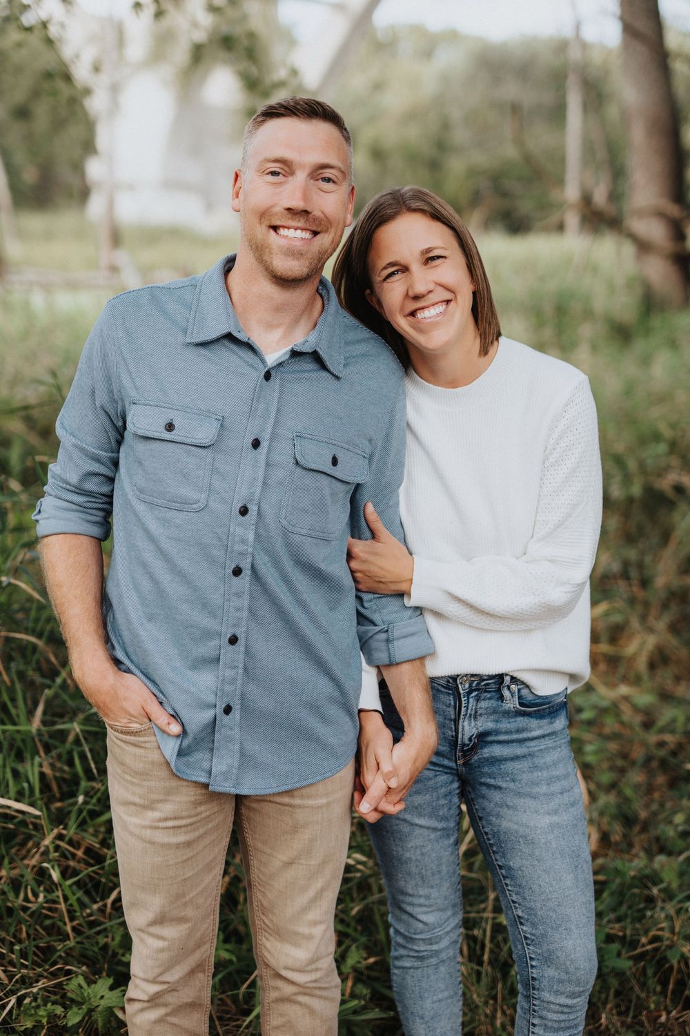 A casual outdoor portrait of a couple in denim and neutral clothing standing together in a natural setting.