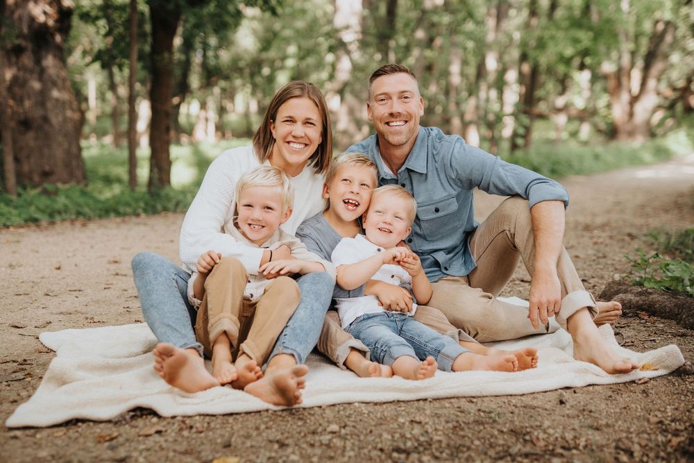 A family wearing coordinated blue and beige outfits poses together on a blanket in a wooded outdoor setting.