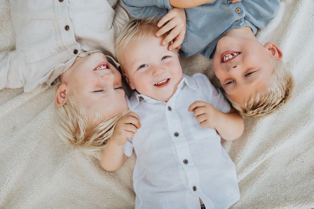 Three young children with light hair lay together on light colored bedding while smiling and laughing.