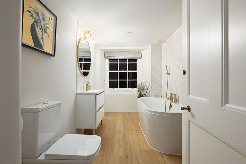 Modern white bathroom interior with freestanding tub, floating vanity, and light wood flooring illuminated by wall sconces.