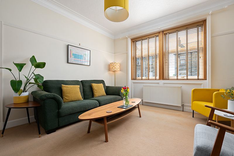 A bright modern living room with green sofa, yellow accents, and hardwood flooring features large windows and plants.