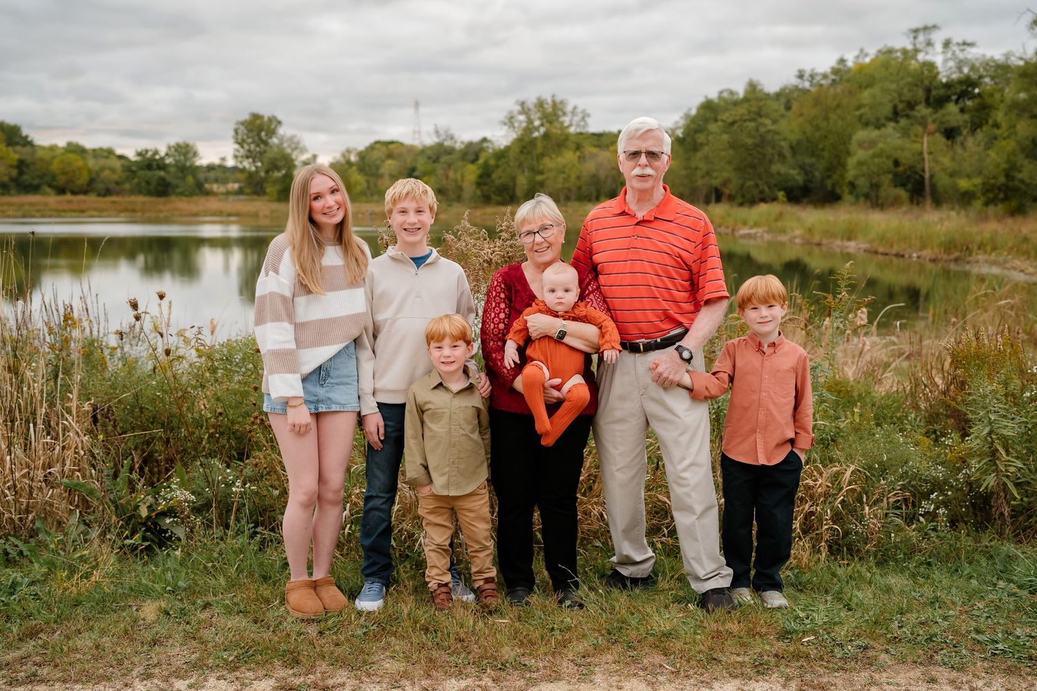 A family group poses together outdoors by a pond surrounded by autumn foliage and tall grass.