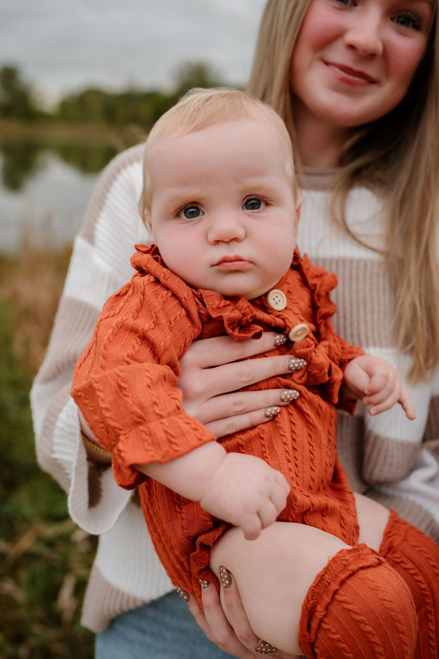 Baby wearing a rust-colored knit sweater being held against an outdoor autumn background.