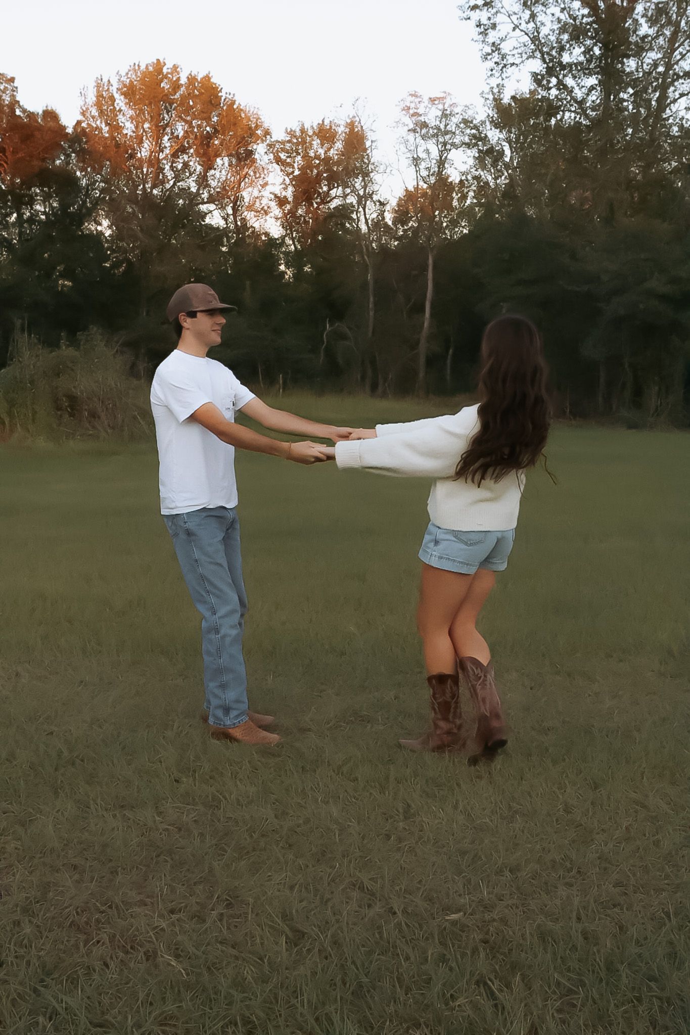 Couple in white shirts and denim playfully spinning in a grassy field.