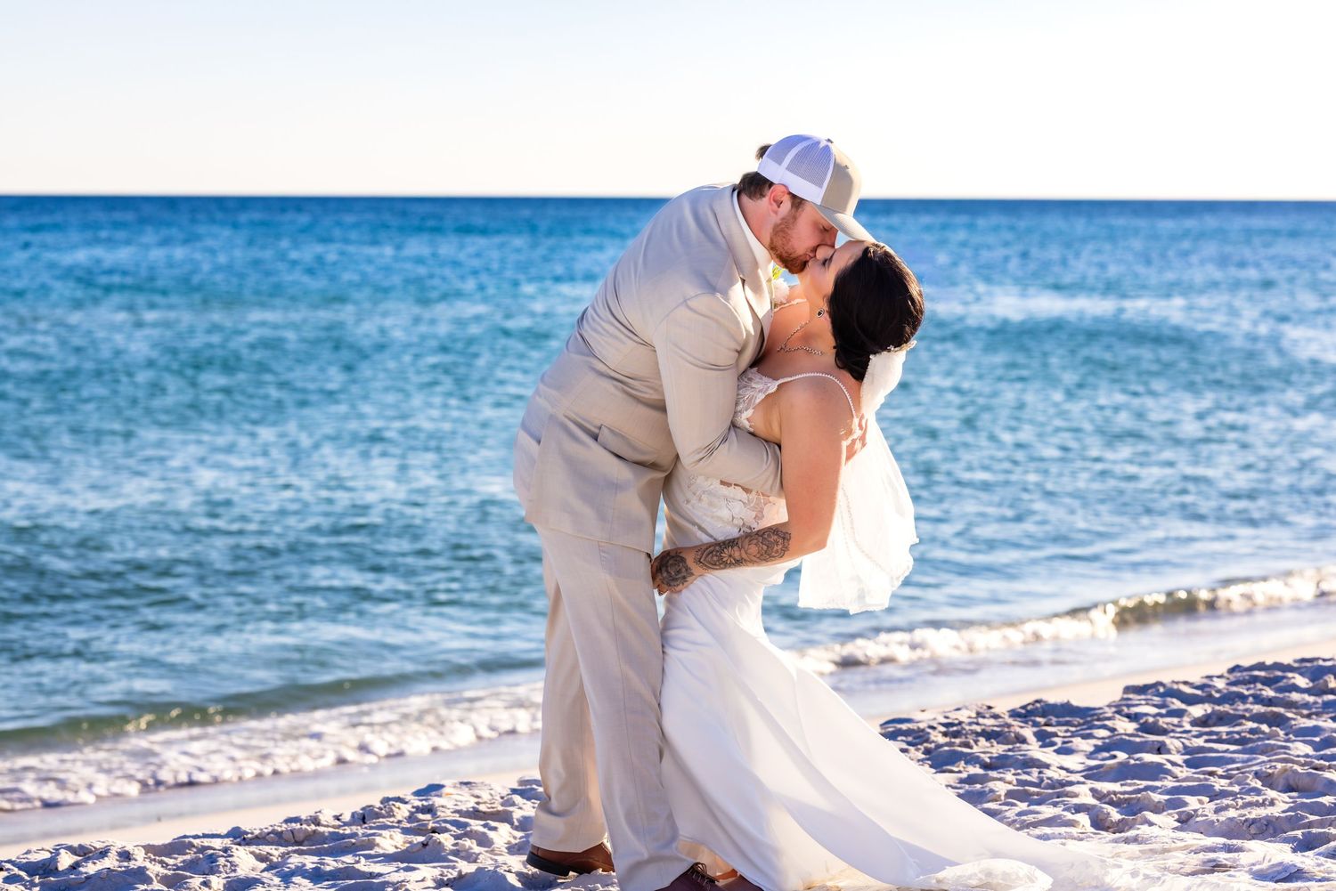 A romantic beach wedding scene with a couple sharing a kiss by the ocean on a sunny day.