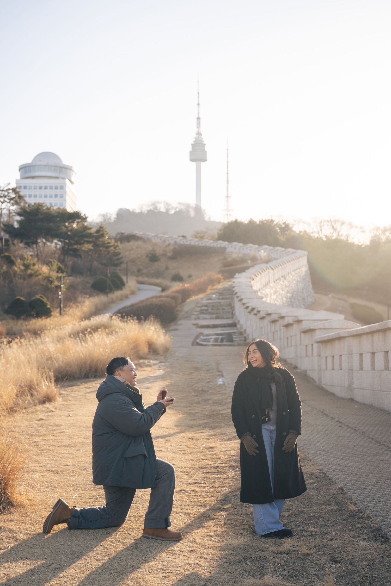 Two people sit together on a hillside path at sunset with a city skyline visible in the distance.