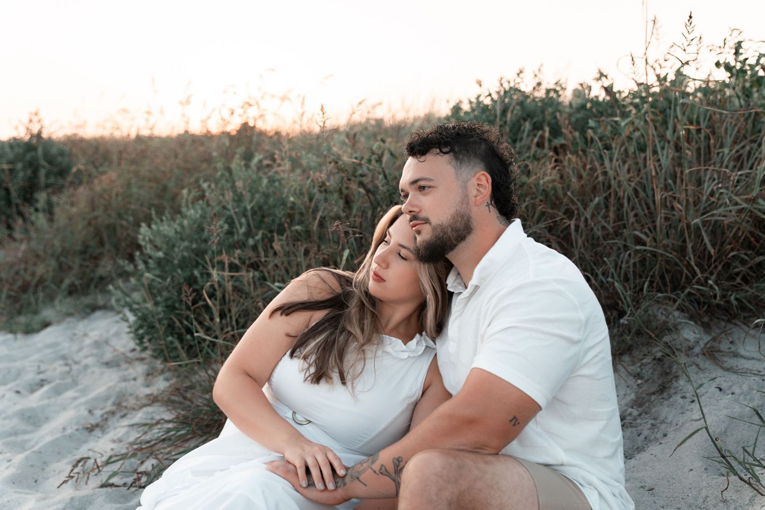 A couple shares an intimate moment while sitting together on sandy dunes at sunset.
