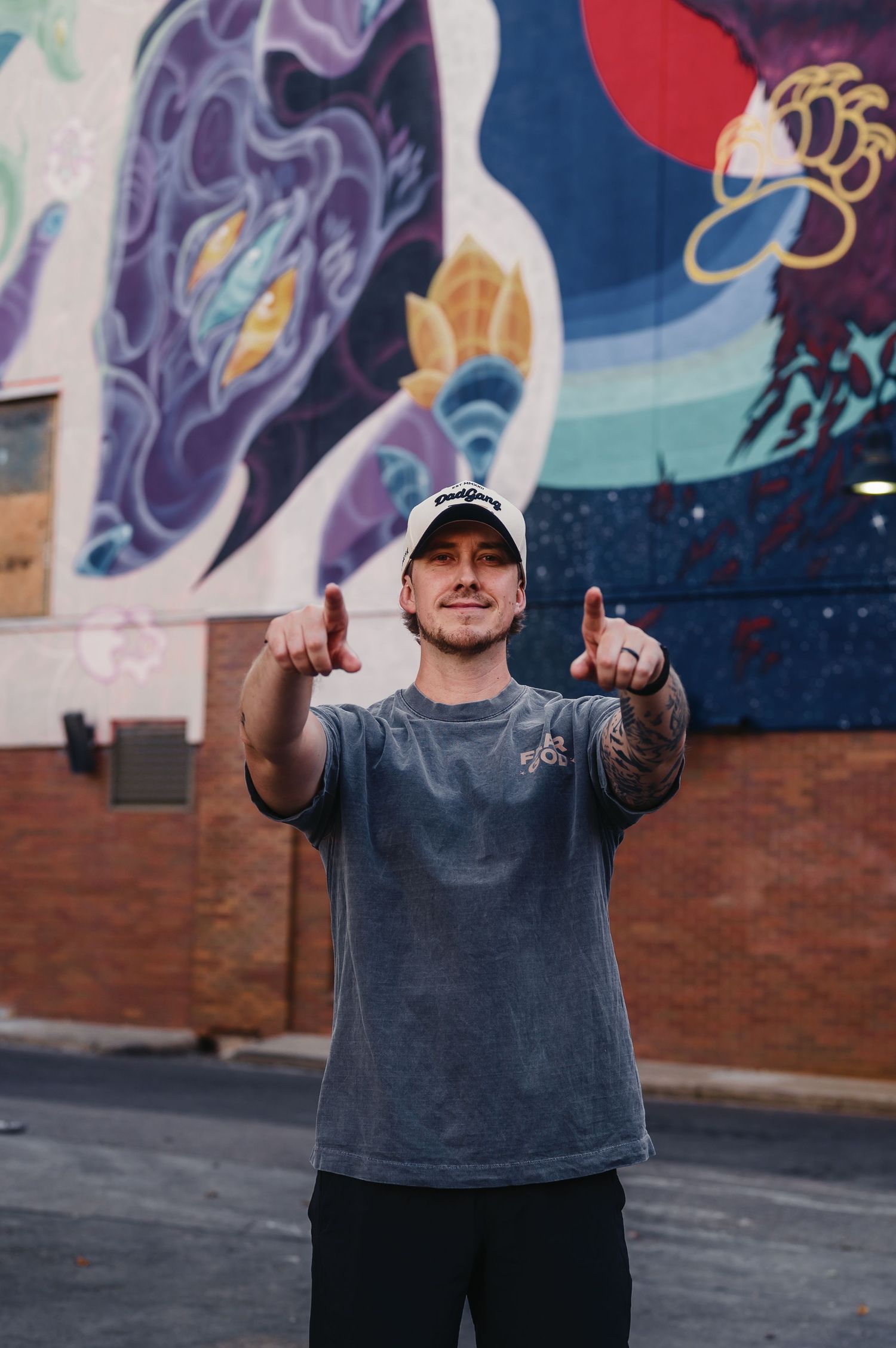 Person in grey t-shirt and black cap gestures against colorful graffiti wall background in urban setting.