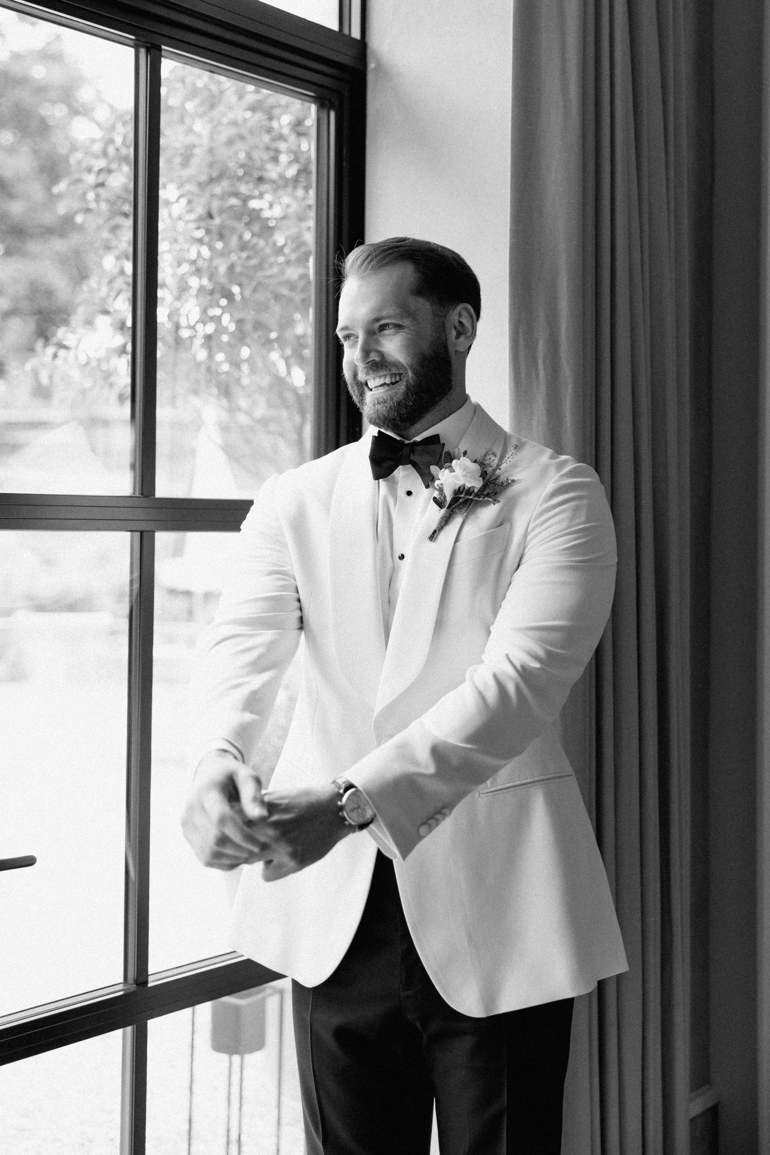 Groom in white tuxedo jacket smiling and getting ready by window.