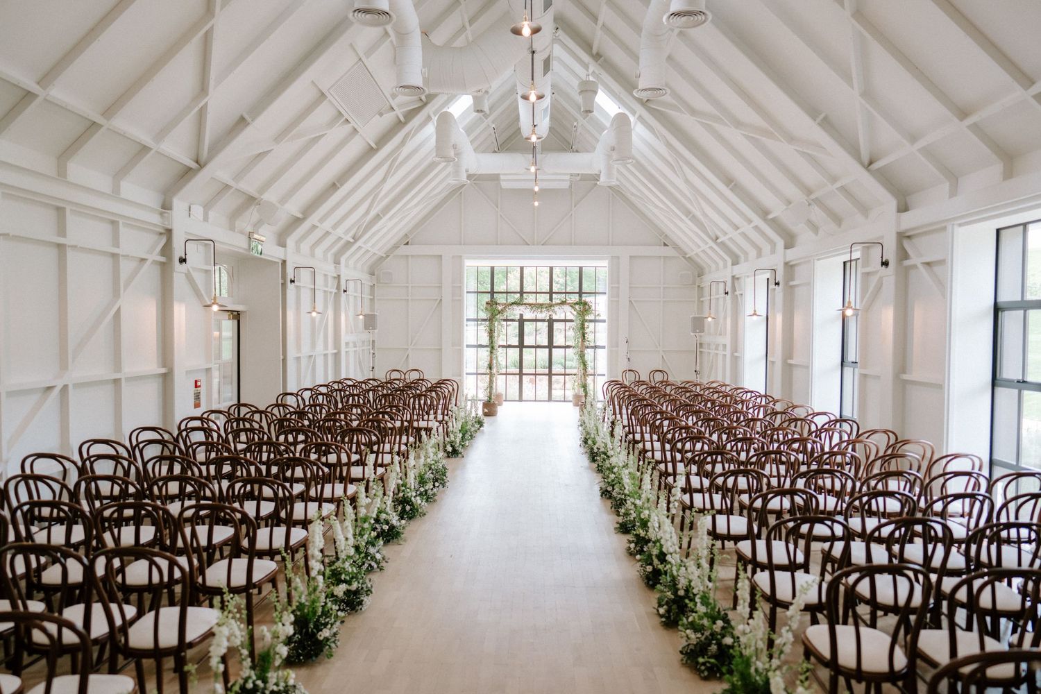 Elegant white wedding venue with wooden chairs and vaulted ceiling decorated with greenery.