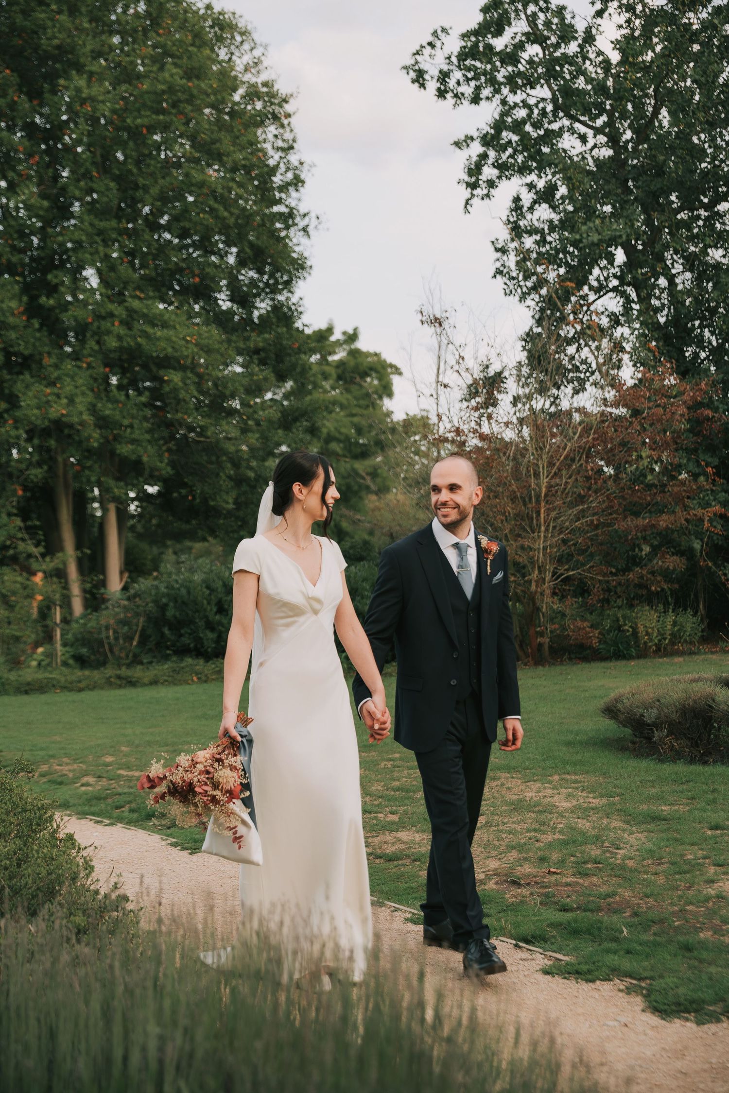 Elegant couple walks garden path carrying autumn-toned bouquet during wedding photoshoot.