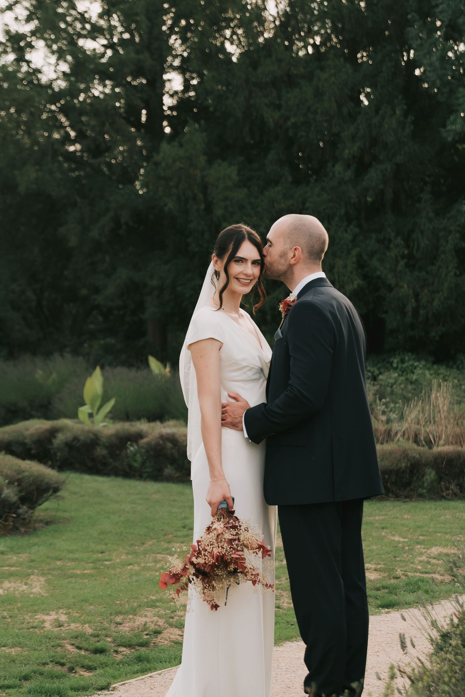 Wedding couple shares intimate moment in garden setting with dried flower bouquet at sunset.