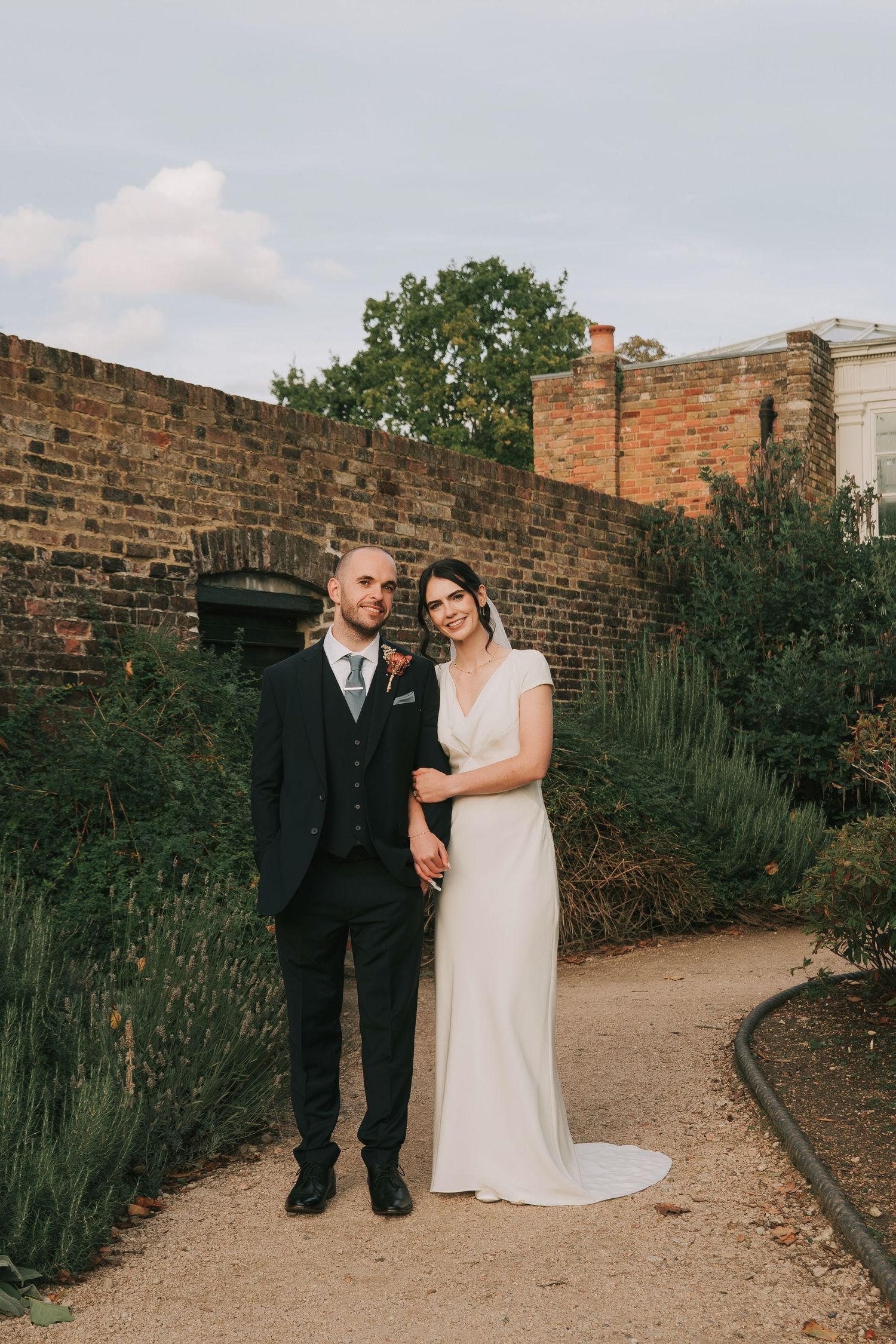 A wedding couple poses along a brick wall pathway with greenery in an outdoor garden setting.
