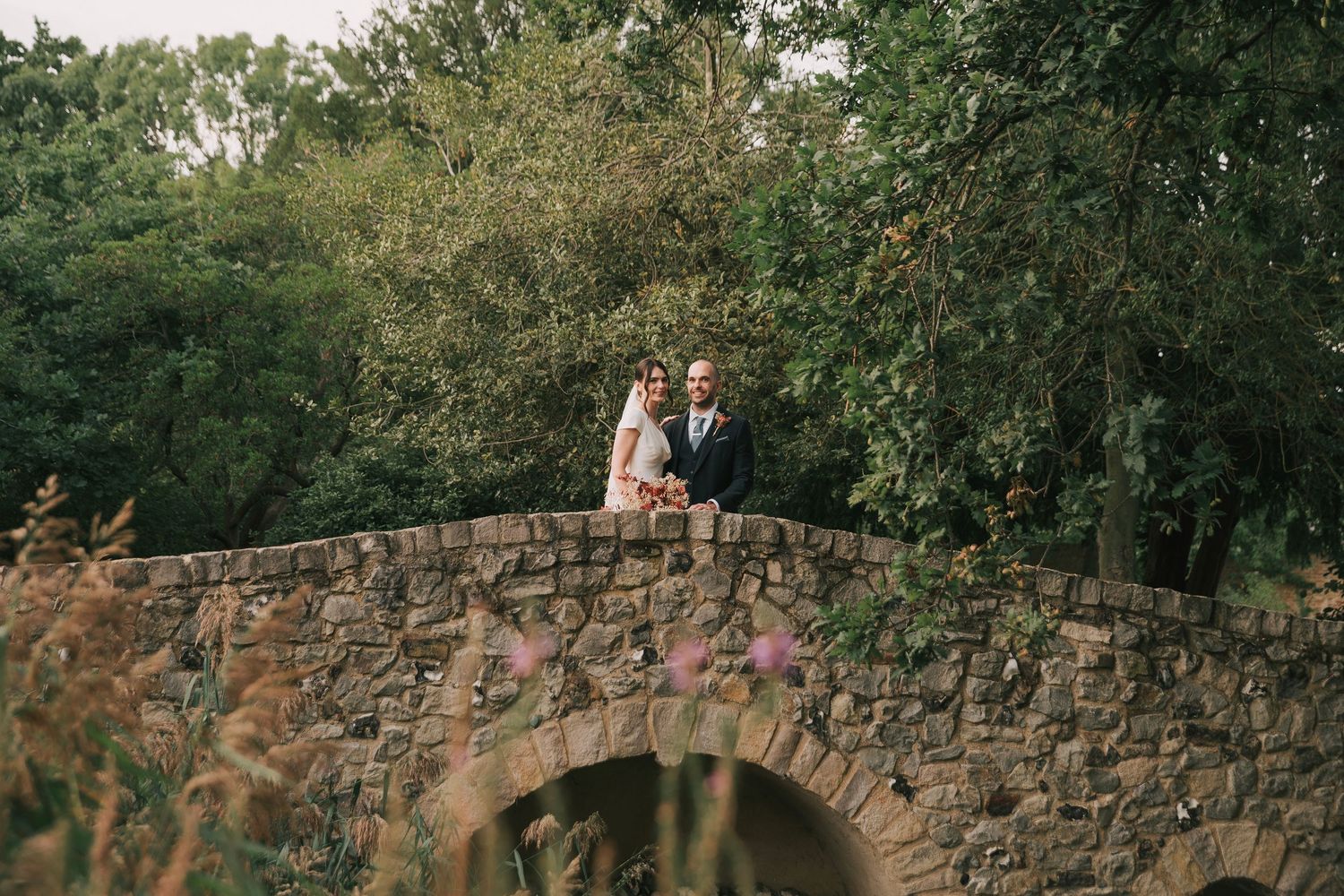 Wedding couple stands on rustic stone bridge surrounded by lush greenery.