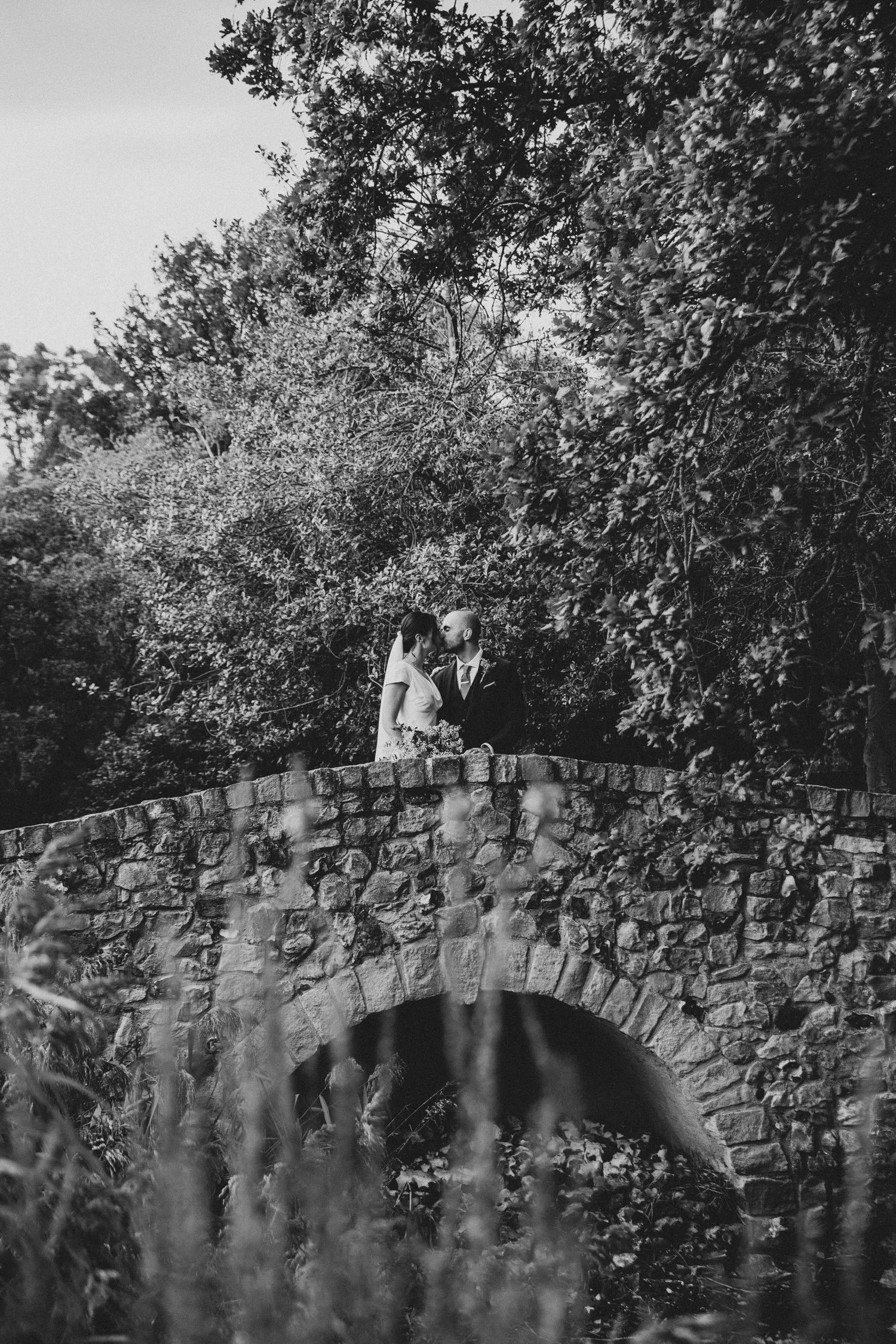 A black and white romantic moment captured on a stone bridge surrounded by lush trees.