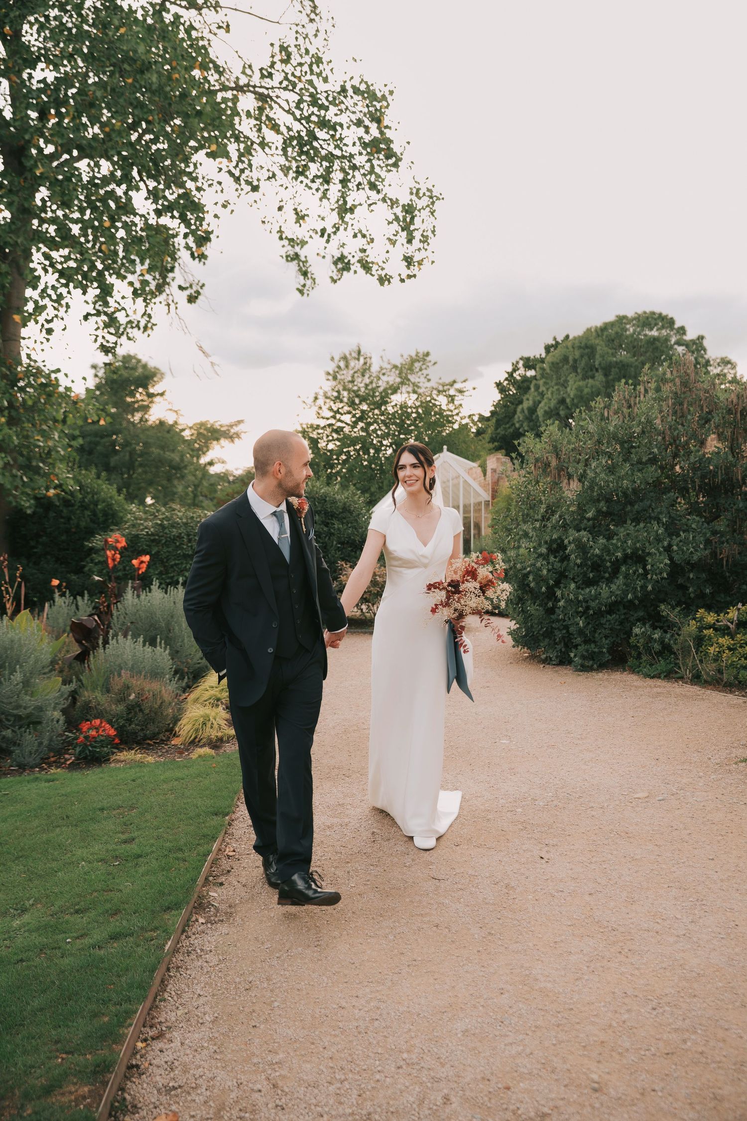 Wedding couple walks along a garden path with trees and landscaping on a cloudy day.