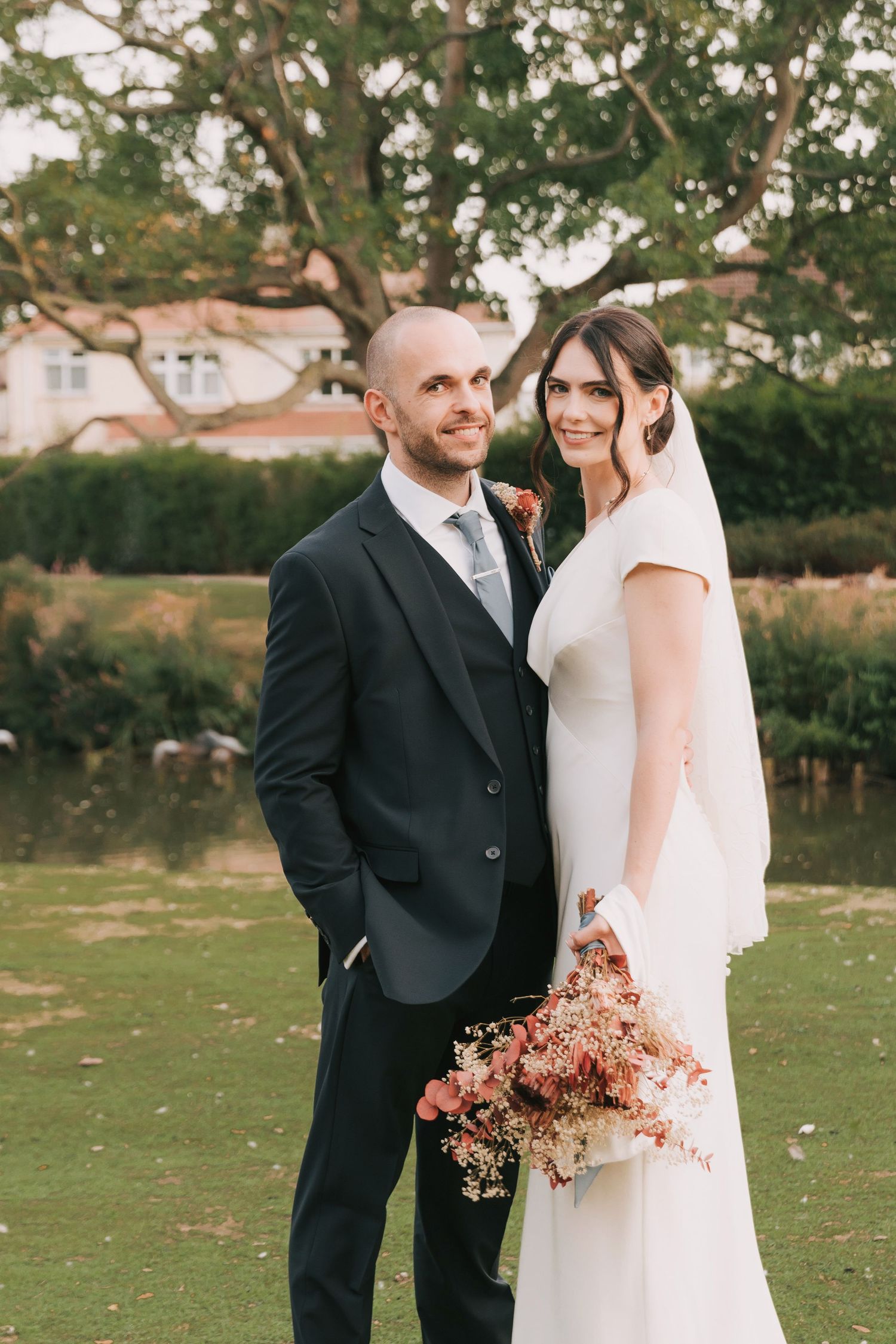 Elegant couple in wedding attire posing together on manicured lawn with autumn flowers.