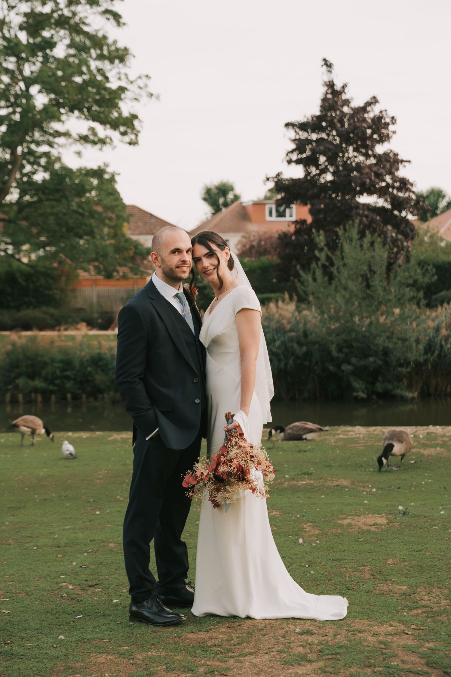 Wedding couple embraces on a green lawn with trees and autumn foliage in the background.