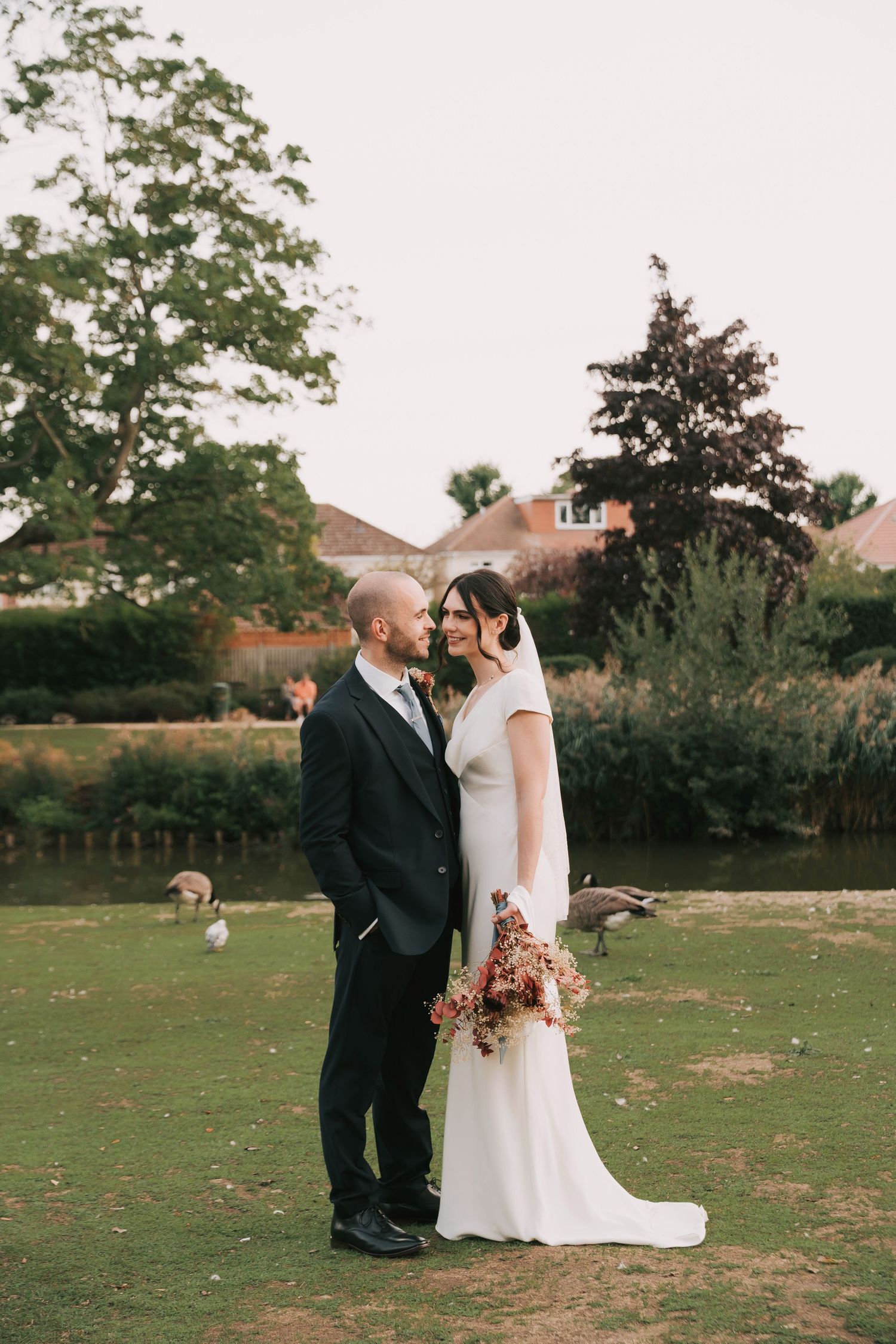 Romantic wedding couple poses together on a lawn with autumn trees in the background.