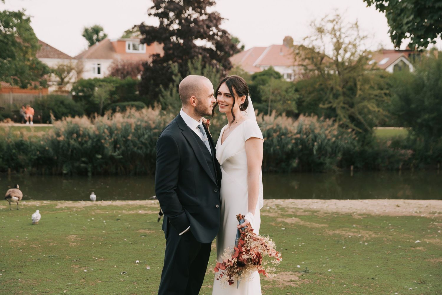 Wedding couple shares tender moment by pond with autumn garden backdrop.
