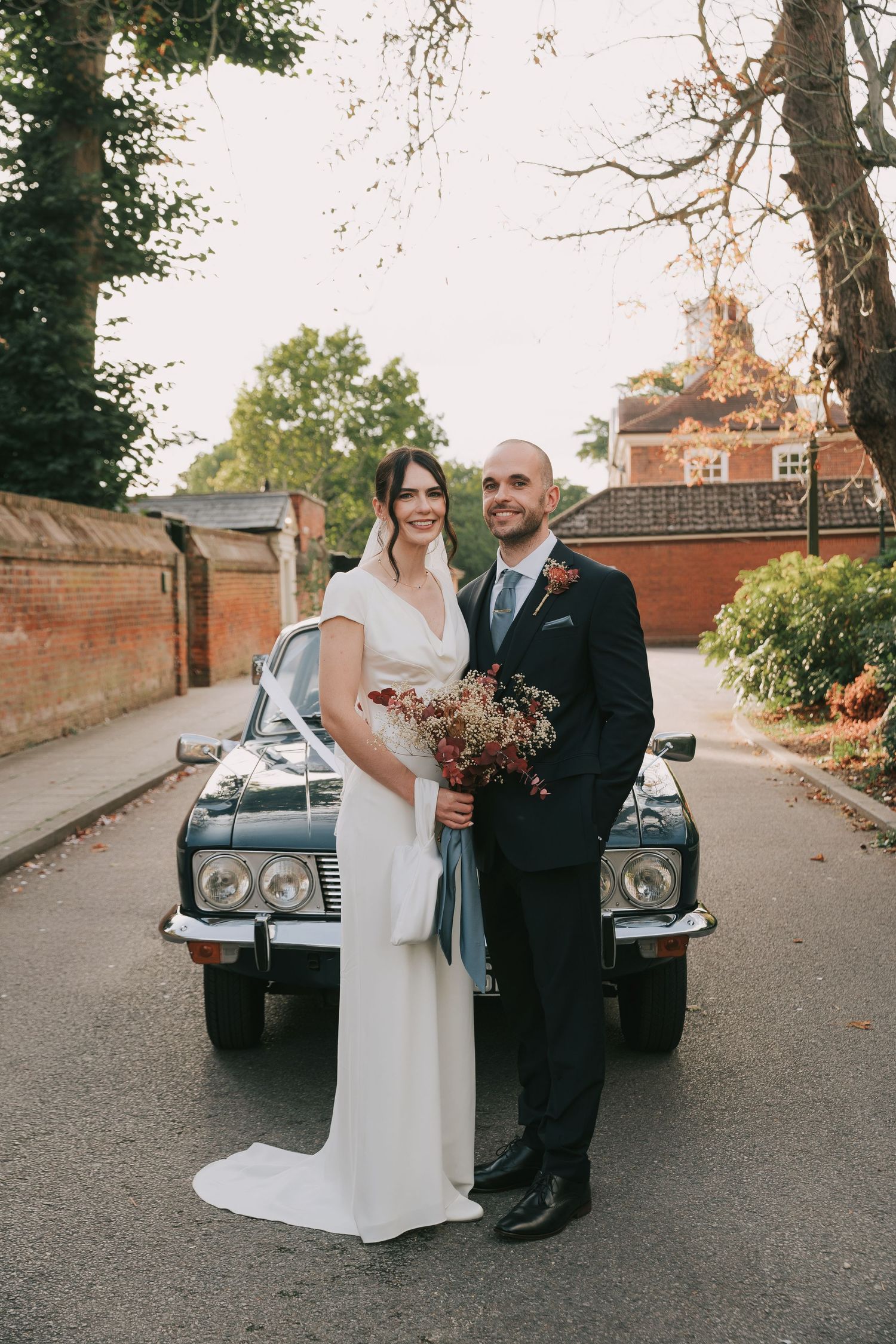 A classic black vintage car serves as an elegant backdrop for a wedding photo on a tree-lined path.