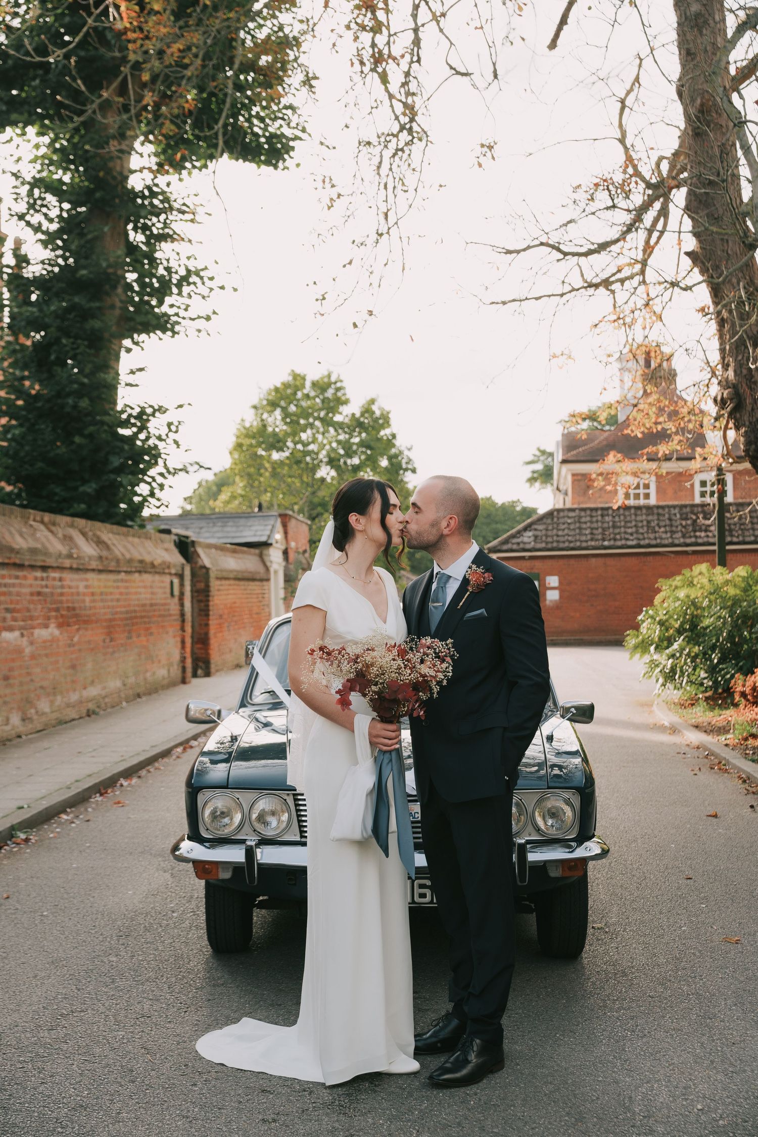 Newlyweds pose with vintage car on brick-lined street with autumn foliage and historic building in background.