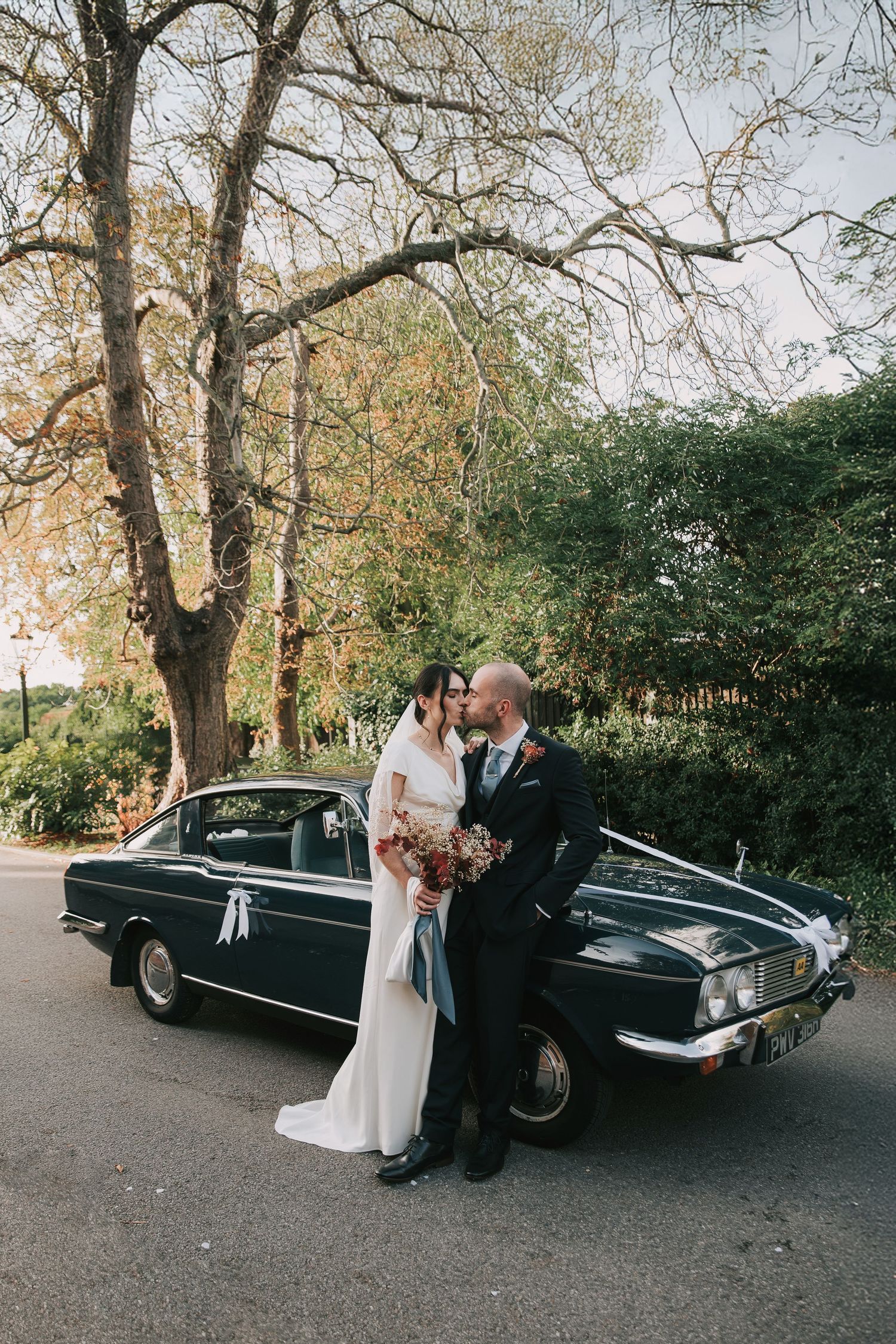 An elegant vintage black car with white ribbons parked beneath autumn trees creates a classic wedding scene.