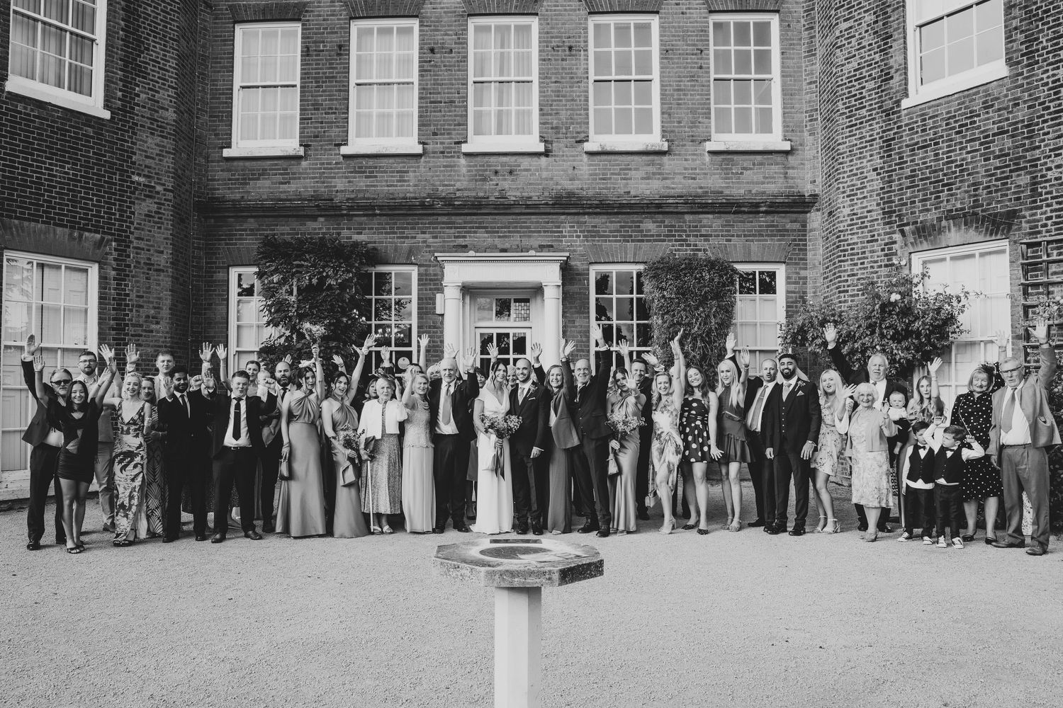 Large group of wedding guests lined up for a formal photo in front of a historic brick building.