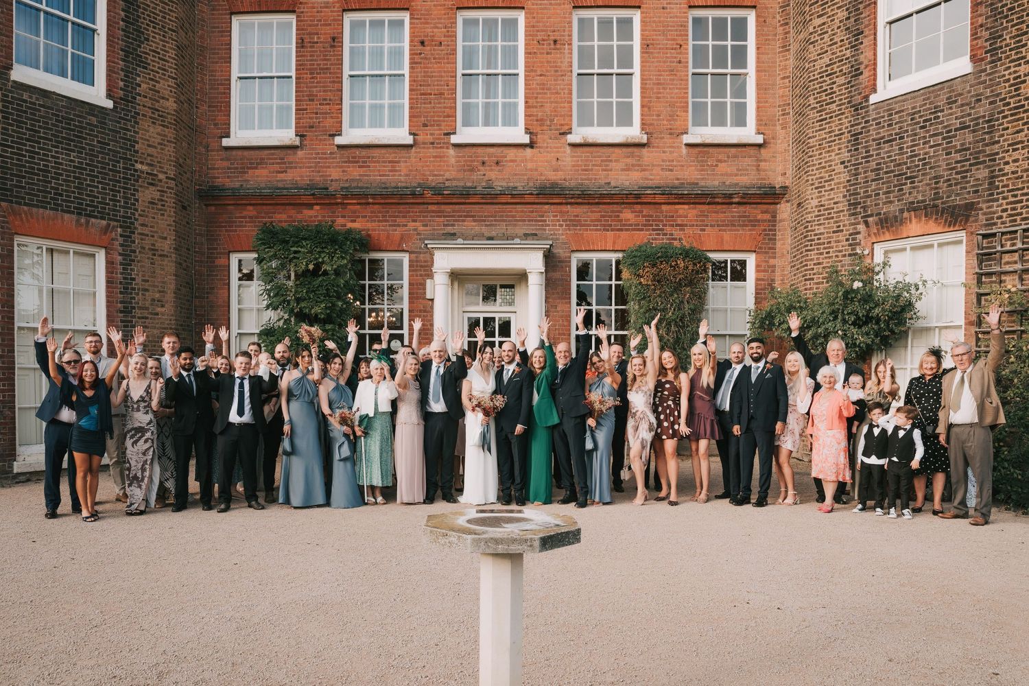 Wedding party and guests gathered for a panoramic group photo outside a traditional brick mansion.
