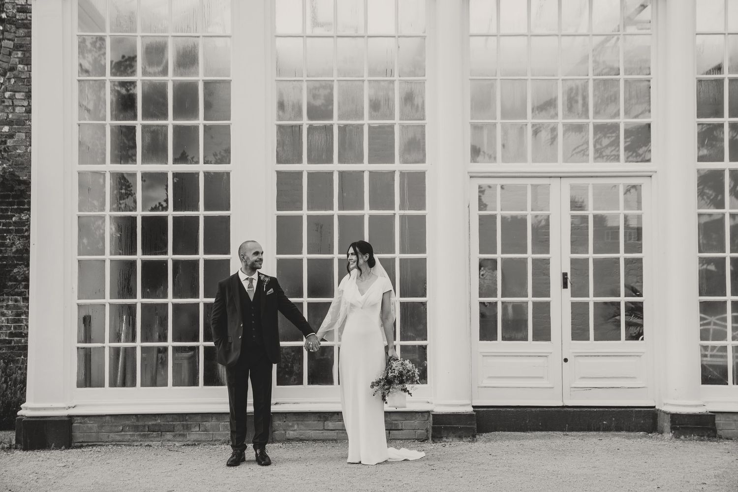 Elegant couple poses in front of a white conservatory with floor-to-ceiling windows.