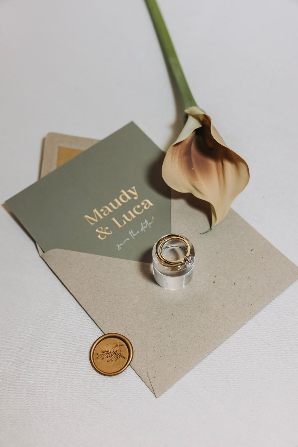 Wedding rings and invitation cards in sage green and beige tones arranged in a minimalist flat lay composition.
